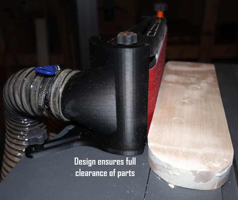 Pu&ograve; includere: Un sistema di raccolta della polvere nero con un tubo collegato a una levigatrice. La levigatrice ha una cinghia abrasiva rossa ed &egrave; fissata a una base nera. Il testo "Design ensures full clearance of parts" &egrave; visibile sotto la levigatrice.
