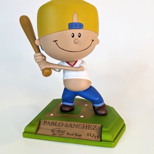 May include: A yellow-haired figurine of a baseball player wearing a blue cap, white shirt, and blue pants. The figurine is holding a wooden bat and is standing on a green base with a wooden plaque that reads "PABLO SANCHEZ TC Print Shop 11/15".