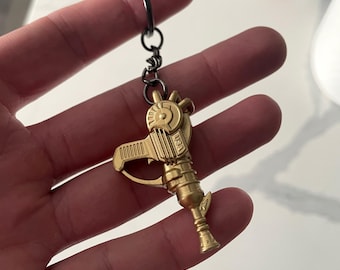 Puede incluir: Un llavero de metal dorado con forma de pistola de rayos retro. El diseño presenta un mango con estrías verticales, un componente lateral circular y una punta de cañón acampanada. Las letras USA están grabadas en el lateral del cuerpo de la pistola. Está unido a una anilla de metal oscuro.
