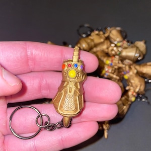 May include: A gold-colored keychain featuring a miniature replica of the Infinity Gauntlet. The gauntlet has six colored gems and intricate detailing. A silver chain and keyring are attached. Another gold-colored figure is visible in the background.