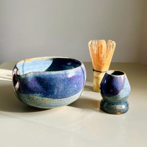 May include: A ceramic matcha bowl with a blue and purple speckled glaze featuring a side pouring spout, accompanied by a bamboo matcha whisk and a matching ceramic whisk holder in a coordinating blue and purple speckled glaze finish.