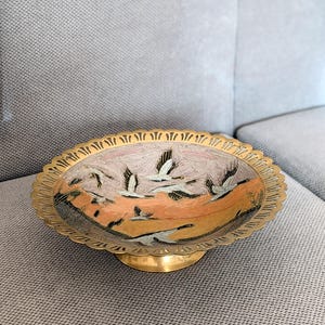 Pode incluir: Uma tigela decorativa de latão com borda ondulada e base de pedestal. O interior da tigela apresenta uma paisagem pintada com pássaros voando contra um fundo de tons laranja e cinza. A tigela é uma peça vintage.