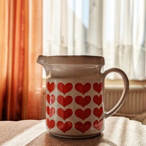 May include: A white ceramic pitcher with a handle and a spout. The pitcher is decorated with a pattern of red hearts. The background includes curtains and natural light.
