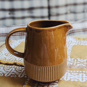 May include: A brown ceramic jug with a handle and a spout. The bottom third of the jug has vertical ribbed detailing. The jug is set on a yellow patterned surface.
