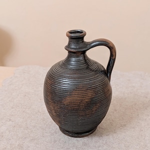 May include: A dark brown, handcrafted ceramic jug with a rounded body and a handle. The jug features horizontal, textured lines around its body and a small opening at the top. The jug is a decorative piece.