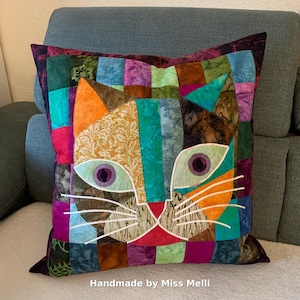 Może przedstawiać: Kolorowa poduszka patchwork z motywem kociej twarzy. Twarz kota wykonana jest z kawałków materiału w odcieniach pomarańczu, turkusu, fioletu i złota, z białymi haftowanymi wąsami. Poduszka jest ręcznie robiona, o czym świadczy napis "Handmade by Miss Melli".