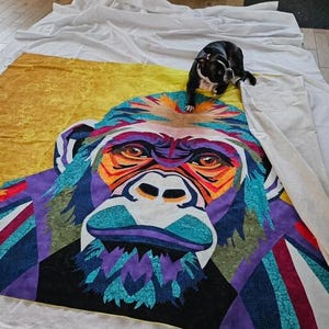 May include: A colorful quilt featuring a close-up of a gorilla's face. The gorilla's face is made up of different colored geometric shapes and patterns. The quilt is laid out on a white sheet.