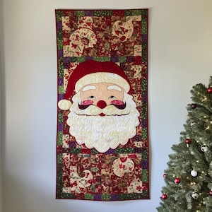 May include: A red, green and white quilted wall hanging featuring a Santa Claus face with a white beard and a red hat. The background is a patchwork of different fabrics with a Christmas theme.