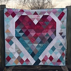 Könnte beinhalten: Eine Steppdecke mit einem großen Herzdesign in Rosatönen, Rot und Türkis. Das Herz besteht aus vielen kleinen Dreiecken und die Steppdecke ist mit einem türkisfarbenen Stoff eingefasst.