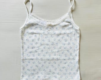 May include: A white ribbed camisole featuring a repeating pattern of small light blue teddy bears. The garment has thin spaghetti straps, a scoop neckline trimmed with delicate white lace, and a small white bow centered at the neckline.