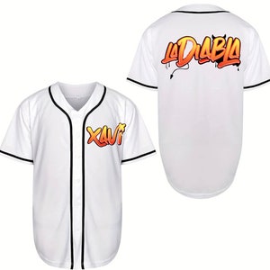 Pode incluir: Camisola de beisebol branca com guarnições pretas. A frente da camisola tem um gráfico da palavra "Xavi" em laranja e amarelo. As costas da camisola têm um gráfico das palavras "La Diablo" em laranja e amarelo.