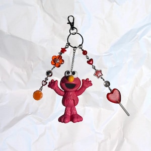 May include: A pink Elmo keychain with a black clasp, adorned with red heart, star, and flower beads, and a lollipop charm. The keychain also features a small orange sphere charm and silver-colored chain accents.