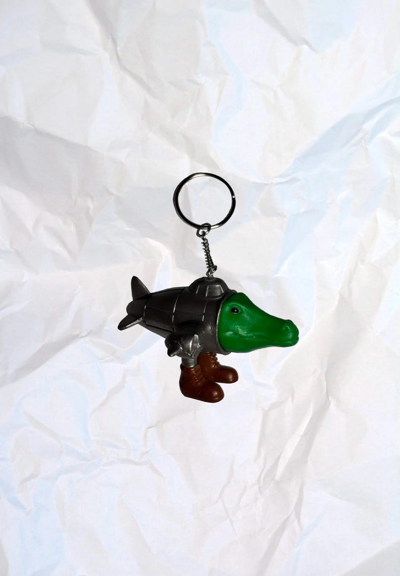Puede incluir: Un llavero novedoso con una cabeza de caim&aacute;n verde y botas marrones unidas a un cuerpo met&aacute;lico similar a un submarino. El llavero tiene un anillo plateado y una peque&ntilde;a cadena.