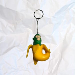 Puede incluir: Un llavero novedoso con un pl&aacute;tano amarillo y una cara de mono verde. El llavero tiene una cadena y un anillo plateados. El fondo es una superficie blanca texturizada.