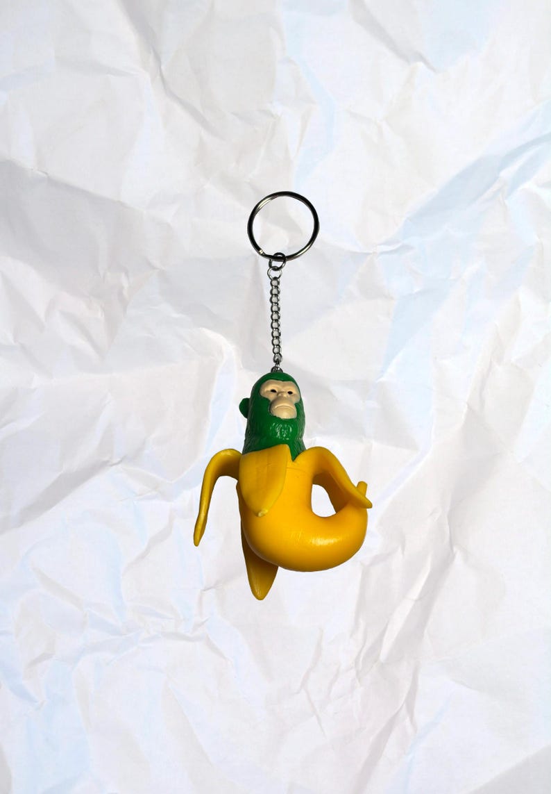 Puede incluir: Un llavero novedoso con un pl&aacute;tano amarillo y una cara de mono verde. El llavero tiene una cadena y un anillo plateados. El fondo es una superficie blanca texturizada.