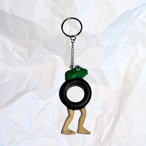 Puede incluir: Un llavero caprichoso con una rana verde posada sobre un neum&aacute;tico negro, con patas color beige que se extienden por debajo. El llavero incluye un anillo y una cadena de metal. La rana tiene un solo ojo visible. El dise&ntilde;o general es l&uacute;dico y &uacute;nico.