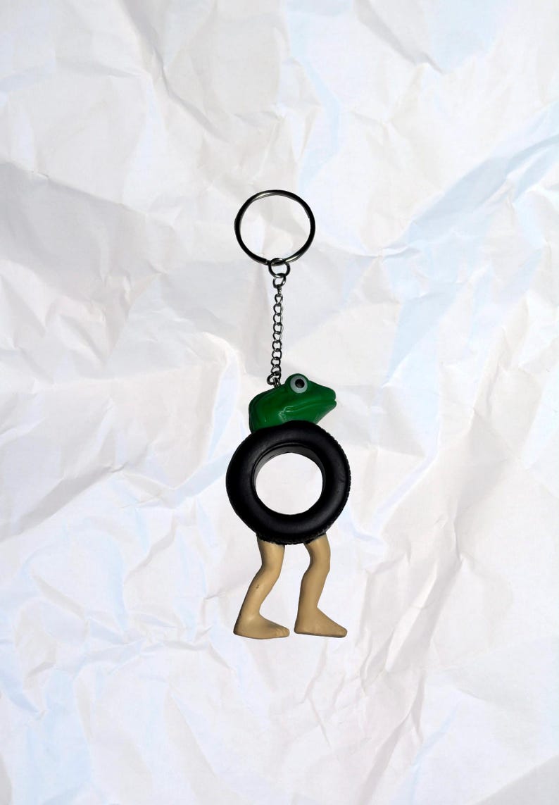 Puede incluir: Un llavero caprichoso con una rana verde posada sobre un neum&aacute;tico negro, con patas color beige que se extienden por debajo. El llavero incluye un anillo y una cadena de metal. La rana tiene un solo ojo visible. El dise&ntilde;o general es l&uacute;dico y &uacute;nico.