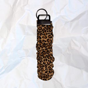 May include: A leopard print keychain wristlet with a black metal ring and a black leather strap. The wristlet is brown and black with a repeating spotted pattern. The background is white.