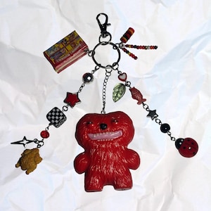 May include: A quirky keychain with a red, furry monster charm and a smiling face. The keychain has a selection of dangling charms, including a miniature box of Barnum's Animals crackers, stars, a ladybird, and beads. Set against a white, textured backdrop.