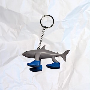 Può includere: Un portachiavi a forma di squalo grigio con scarpe blu. Lo squalo ha una catena e un anello di metallo per attaccarlo alle chiavi o a una borsa. Un oggetto divertente.