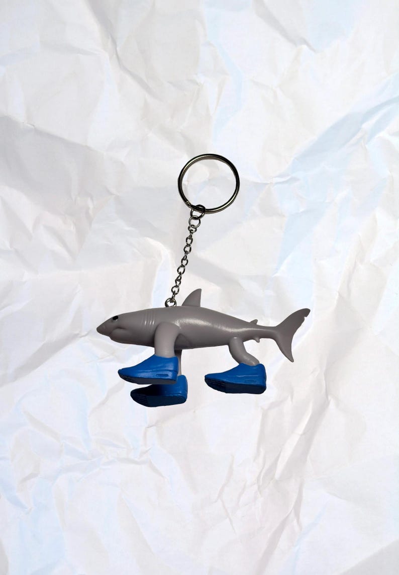Puede incluir: Un llavero de tibur&oacute;n gris con zapatos azules. El tibur&oacute;n tiene una cadena y un anillo de metal para sujetar a llaves o una bolsa. Un art&iacute;culo divertido y original.