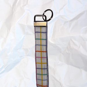 May include: A gray fabric keychain wristlet with a colorful plaid pattern. The wristlet has a gold-colored metal accent and a black metal key ring. The plaid design features yellow, green, purple, and orange lines.