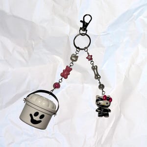 Puede incluir: Un llavero decorativo con un cubo blanco con una cara sonriente, un personaje de Hello Kitty y varios abalorios. Los abalorios incluyen un oso de peluche rosa, un hueso y cuentas en forma de estrella. Tiene un cierre negro.