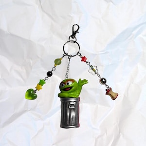 Könnte beinhalten: Ein skurriler Schlüsselanhänger mit einer grünen Figur, die aus einem silbernen Mülleimer kommt. Der Schlüsselanhänger enthält dekorative Charms mit Sternen, Herzen und Perlen. Ein lustiges Accessoire für Taschen oder Schlüssel.