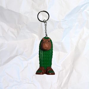 Puede incluir: Un llavero novedoso con una figura caprichosa verde, parecida a un cactus, con cabeza y pies de hipop&oacute;tamo marr&oacute;n. El llavero tiene un anillo y una cadena de color plateado. La figura mide aproximadamente 7,6 cm de alto.