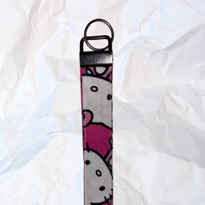 May include: A pink and white fabric keychain wristlet with a repeating cartoon cat face pattern. It has a black metal ring and a black metal clasp.