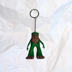 Puede incluir: Un llavero novedoso con una figura verde con cara y pies marrones. La figura tiene los brazos extendidos y est&aacute; unida a una cadena y un llavero de metal. El fondo es una superficie blanca texturizada.