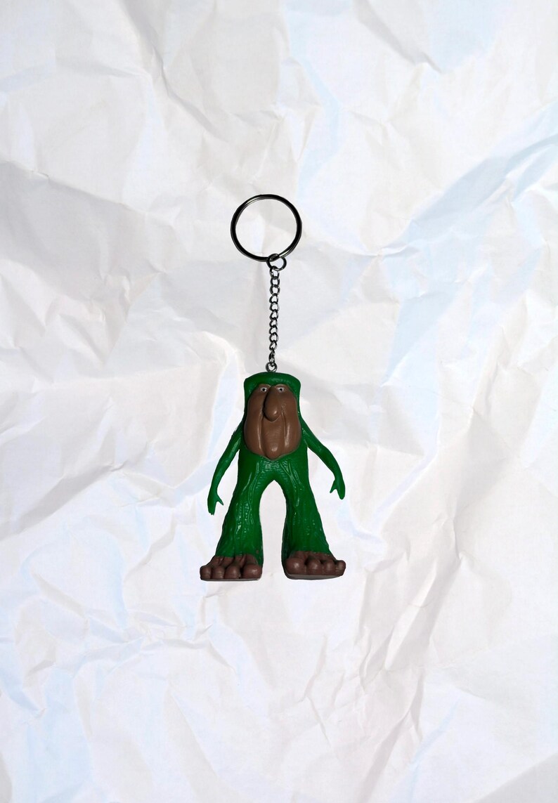 Puede incluir: Un llavero novedoso con una figura verde con cara y pies marrones. La figura tiene los brazos extendidos y est&aacute; unida a una cadena y un llavero de metal. El fondo es una superficie blanca texturizada.