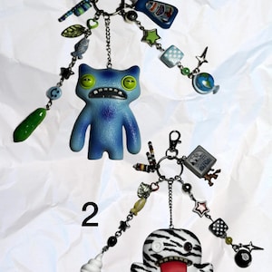 May include: Two keyrings with decorative charms. One features a blue monster figure with green eyes, and the other has a zebra-striped monster. Charms include stars, a globe, a pickle, and an ice cream cone.