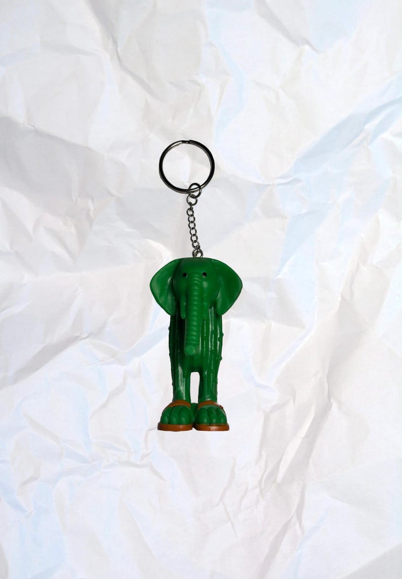Puede incluir: Llavero de elefante verde con cadena y anilla plateadas. El elefante tiene patas marrones y un cuerpo texturizado. Un accesorio divertido para llaves o bolsos.