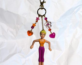 Könnte beinhalten: Ein Schlüsselanhänger mit einer Puppe mit blonden Haaren, einem orangefarbenen Oberteil und einer lila Hose. Der Schlüsselanhänger hat einen goldenen Verschluss und ist mit rosa und orangefarbenen Perlen, Sternen, Herzen und einem Blumenanhänger verziert.