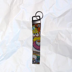 Pode incluir: Um porta-chaves de tecido colorido com fecho de metal e argola. O tecido apresenta um padrão repetido de personagens de desenhos animados em amarelo, rosa, laranja e azul. A palavra "DOSA" é visível no tecido.