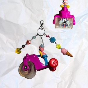 May include: A selection of vintage keyrings with cartoon characters and colourful charms. One keyring has a pink tricycle with a character riding it, and another has a pink roller with a character inside. Charms include stars, hearts, and an ice cream cone.