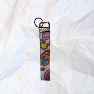 Pode incluir: Um porta-chaves com um anel de metal preto e uma correia de tecido estampado. A correia apresenta personagens de desenhos animados em rosa, amarelo, azul e verde sobre um fundo branco. O porta-chaves é retangular e tem um fecho de metal.