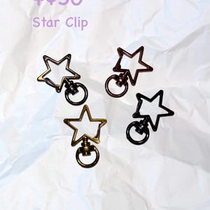 Puede incluir: Cuatro clips en forma de estrella en oro, bronce y negro. Cada clip tiene un anillo adjunto debajo de la estrella. El texto "Star Clip" es visible en la imagen.