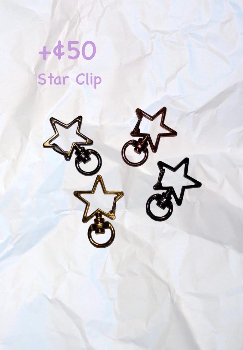 Puede incluir: Cuatro clips en forma de estrella en oro, bronce y negro. Cada clip tiene un anillo adjunto debajo de la estrella. El texto "Star Clip" es visible en la imagen.