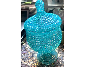May include: A turquoise, bedazzled jar with a lid. The jar is covered in small, iridescent rhinestones, giving it a sparkling appearance. The jar has a rounded base and a decorative top.