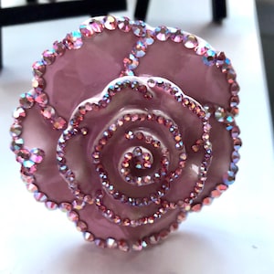 Puede incluir: Un objeto decorativo rosa en forma de rosa con pedrería iridiscente que perfila los pétalos. El centro de la rosa presenta un diseño en espiral, también adornado con pedrería. El objeto probablemente está hecho de resina o un material similar.