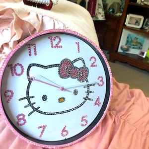 Puede incluir: Un reloj de pared rosa Hello Kitty con pedrería alrededor del borde y una cara de Hello Kitty en el centro. La esfera del reloj es blanca con números y manecillas negros.