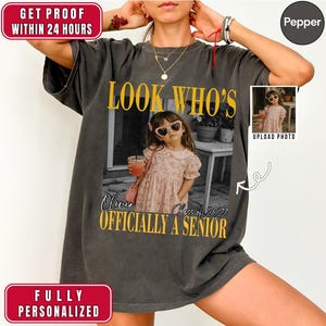 May include: A charcoal grey short-sleeve t-shirt featuring a large central graphic with the text LOOK WHO'S OFFICIALLY A SENIOR in yellow. The graphic includes a black and white photograph of a child in a floral dress and sunglasses.