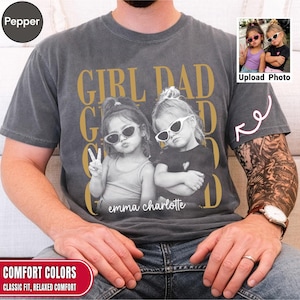 Op de afbeelding: Een antracietgrijs t-shirt met korte mouwen en een grote grafische print met de tekst GIRL DAD in gouden letters. Het ontwerp bevat een zwart-witfoto van twee kinderen met een zonnebril, met de namen emma charlotte eronder gedrukt.