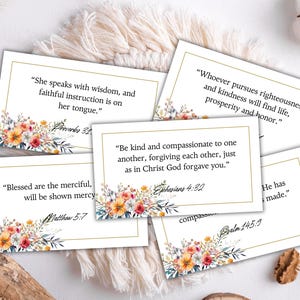 Peut inclure: Plusieurs cartes blanches avec des bordures dorées, chacune présentant un verset biblique en texte noir. Des motifs floraux à l'aquarelle orange, jaune et rose ornent le bas de chaque carte. Les cartes sont disposées sur une surface blanche texturée.