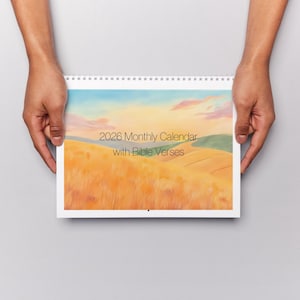 Pode incluir: Calendário mensal de 2026 com encadernação em espiral e uma ilustração de paisagem. A obra de arte apresenta um campo de relva dourada sob um céu azul e laranja, com o texto "2026 Monthly Calendar with Bible Verses". O calendário é segurado por duas mãos.