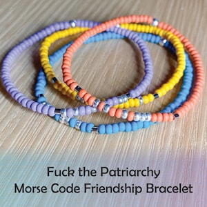 May include: A multi-colored friendship bracelet made of small beads in shades of purple, yellow, orange, and blue. The bracelet has silver beads interspersed throughout. The bracelet is on a wooden surface and the text "Fuck the Patriarchy Morse Code Friendship Bracelet" is below the bracelet.