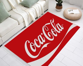 Könnte beinhalten: Ein rechteckiger roter Teppich mit dem Coca-Cola-Logo in Weiß. Der Teppich zeigt den klassischen Coca-Cola-Schriftzug und ein weißes geschwungenes Design am unteren Rand. Der Teppich liegt auf einem weißen Boden in einem Wohnzimmer.