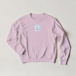 K&ouml;nnte beinhalten: Hellrosa Sweatshirt mit einem kleinen quadratischen Grafik auf der Brust. Die Grafik zeigt ein blau-wei&szlig;es Blumendesign mit einem zentralen Emblem und umliegenden Bl&auml;ttern. Das Sweatshirt hat lange &Auml;rmel und einen Rundhalsausschnitt.